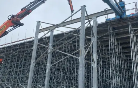 A crane lifting beams onto a large metal bleacher structure under an overcast sky.