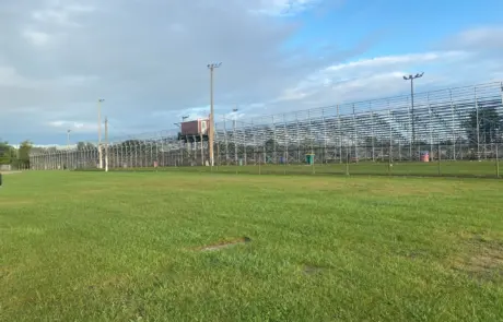 A distant view of a large outdoor bleacher structure behind a chain-link fence on a grassy field.