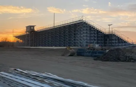 A bleacher structure at sunset with piles of metal beams in the foreground and a clear sky.