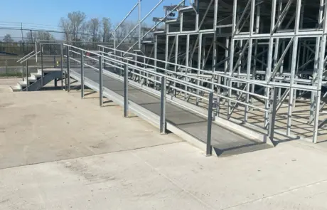 A close-up of a metal bleacher structure with a paved ground and a chain-link fence in the background.